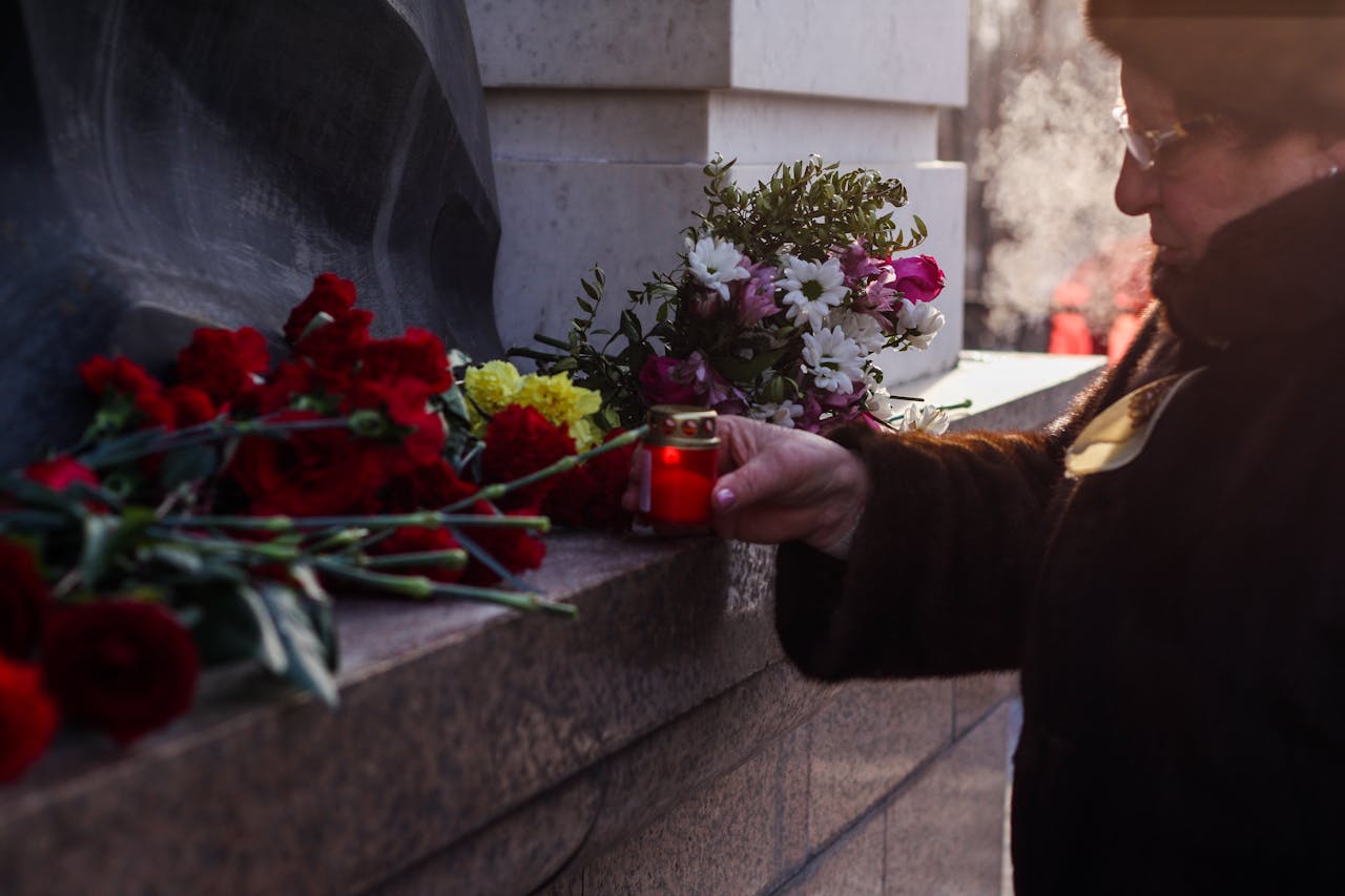 Inicio An elderly woman places flowers and a candle at a cemetery memorial site, reflecting homage and remembrance.