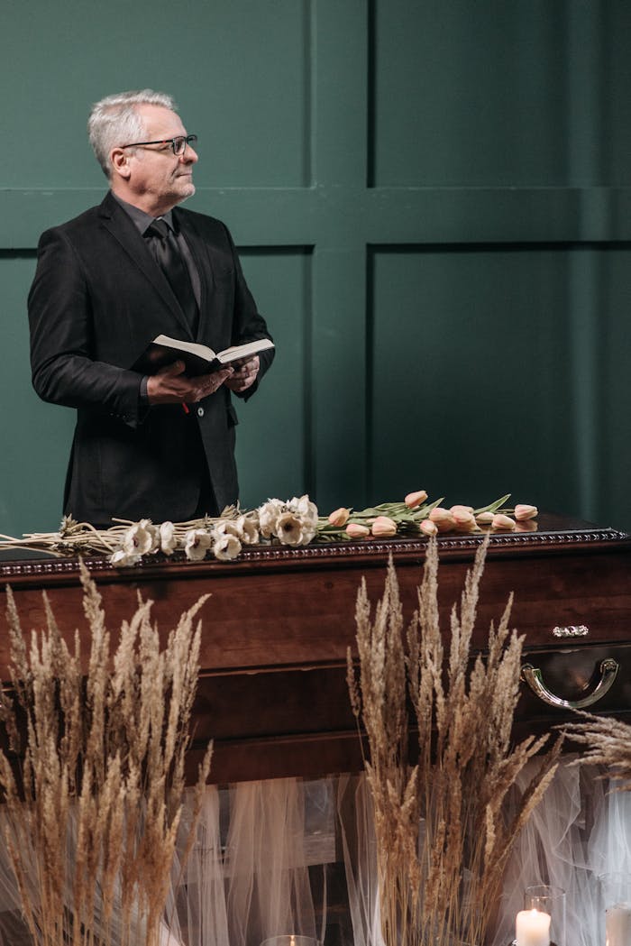 Mature man holding a book beside a coffin with flowers at a funeral service indoors.